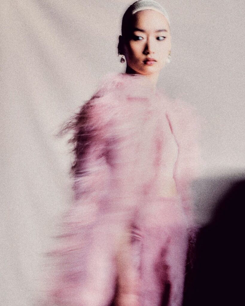 A person in a fuzzy pink outfit stands against a neutral background. The image is intentionally blurred, giving a dreamy, ethereal effect.