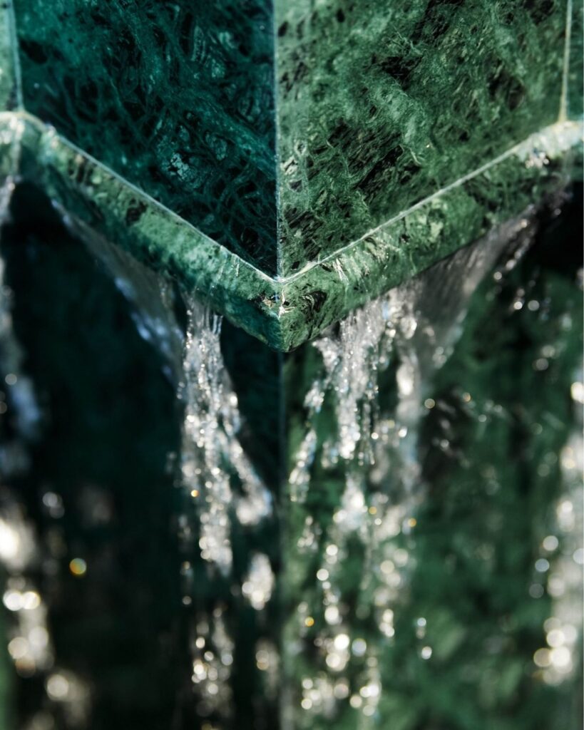 Water cascades down the corners of a textured, green marble structure, creating shimmering flows and reflections on the stone surface