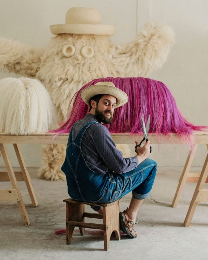 A man in overalls and a hat sits on a stool, holding large scissors. He faces the camera, with long faux hair in white and pink on tables