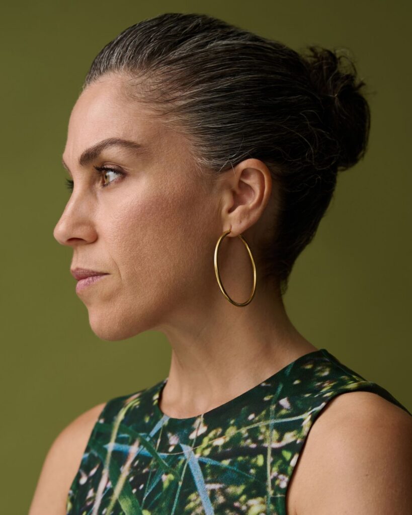 A woman with dark hair in a bun, wearing a sleeveless patterned top and large gold hoop earring, looks to the left against an olive green background