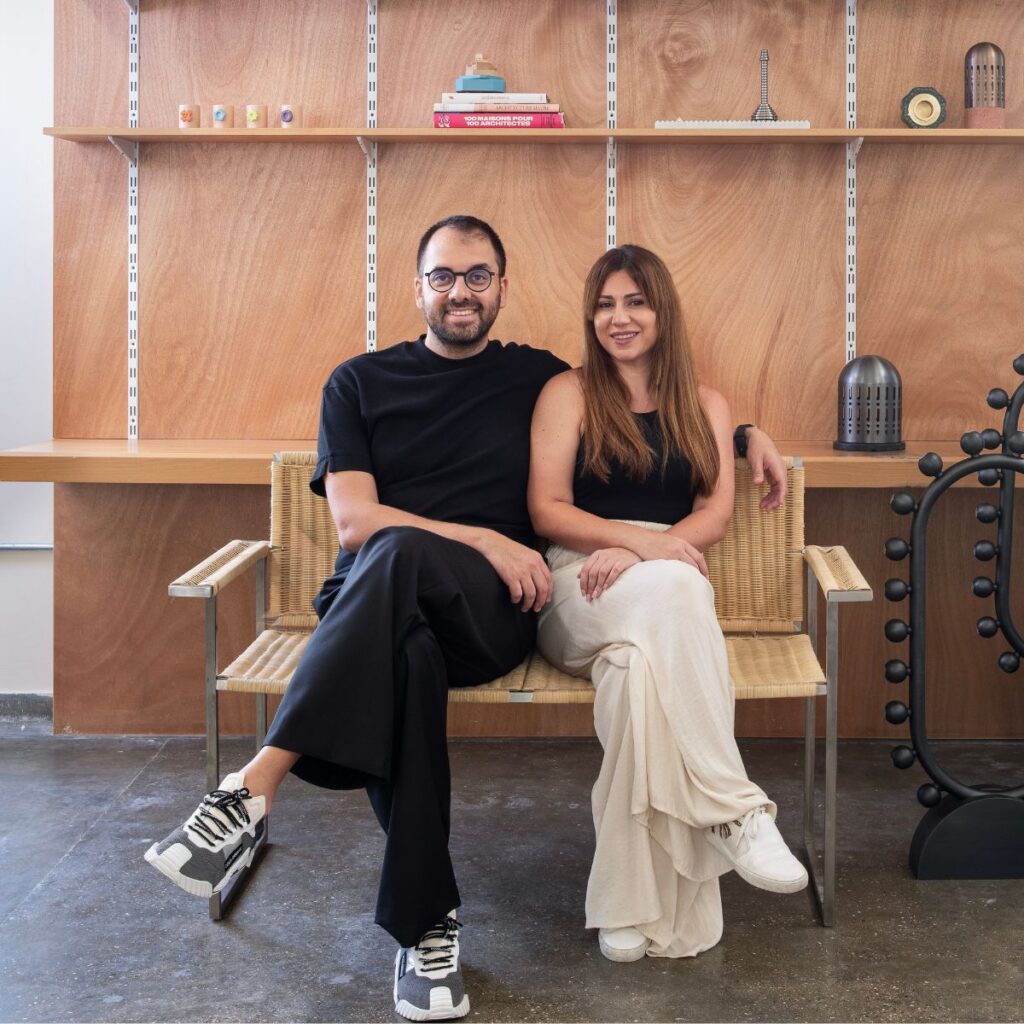 A man and a woman sit on a woven bench in a modern room with wooden shelves, books, and decor. Both are casually dressed in black tops and light pants, smiling at the camera