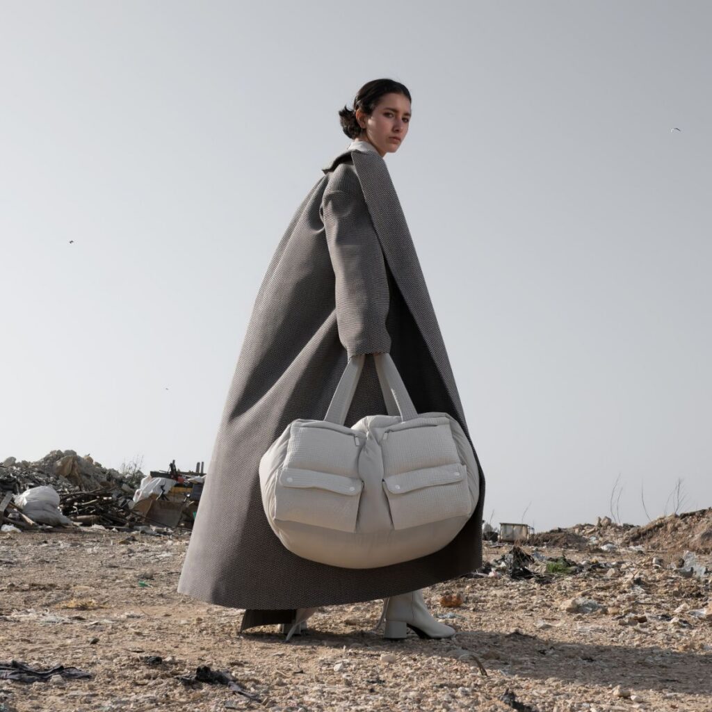 A woman in a long gray coat stands on barren ground holding a large white bag, with debris and a gray sky in the background