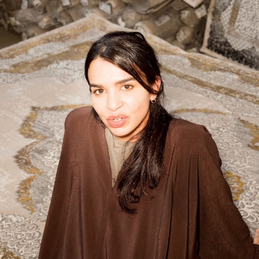 A person with long dark hair wearing a brown outfit stands on patterned rugs, looking up at the camera. Rolls of rug material are stacked in the background, evoking a scene reminiscent of Frida Escobedo’s thoughtful approach to texture and space.