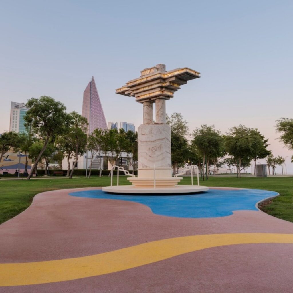 A marble sculpture stands on a circular platform in a park with colorful winding paths. Trees surround the area, and modern tall buildings—reminiscent of Frida Escobedo’s architectural style—are visible in the background under a clear sky.