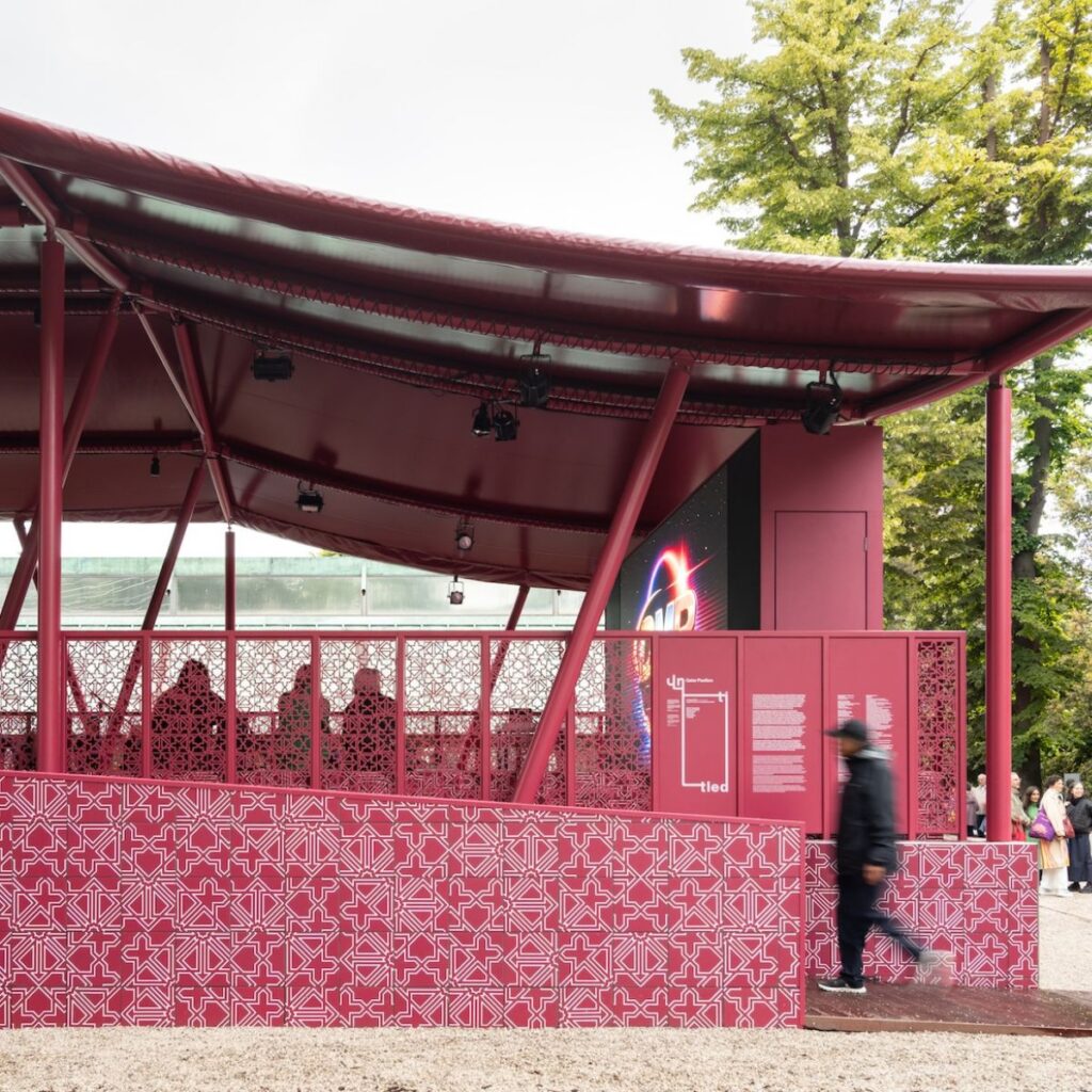 A person walks past a modern pavilion with a geometric patterned pink facade, angled roof, and decorative panels—reminiscent of Msheireb’s contemporary architectural style—surrounded by trees and gravel ground.