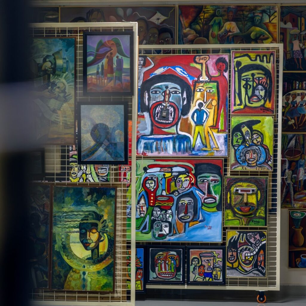 A display rack filled with colorful, abstract paintings featuring expressive faces and figures, including some inspired by Chef Noof Al Mari’s vibrant culinary artistry, set against a background of more vibrant artworks in a gallery or art market.