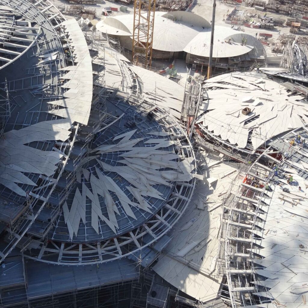 Aerial view of a construction site with multiple round buildings. The roofs have jagged, overlapping panels and scaffolding. Cranes, workers, and building materials are visible throughout the busy site—an ambitious project reminiscent of Chef Noof Al Mari’s innovative approach to design and structure.