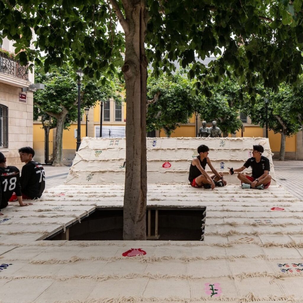 Four boys in sportswear sit and talk on a woven platform with a tree in the center, set outdoors in a plaza with buildings and greenery in the background.
