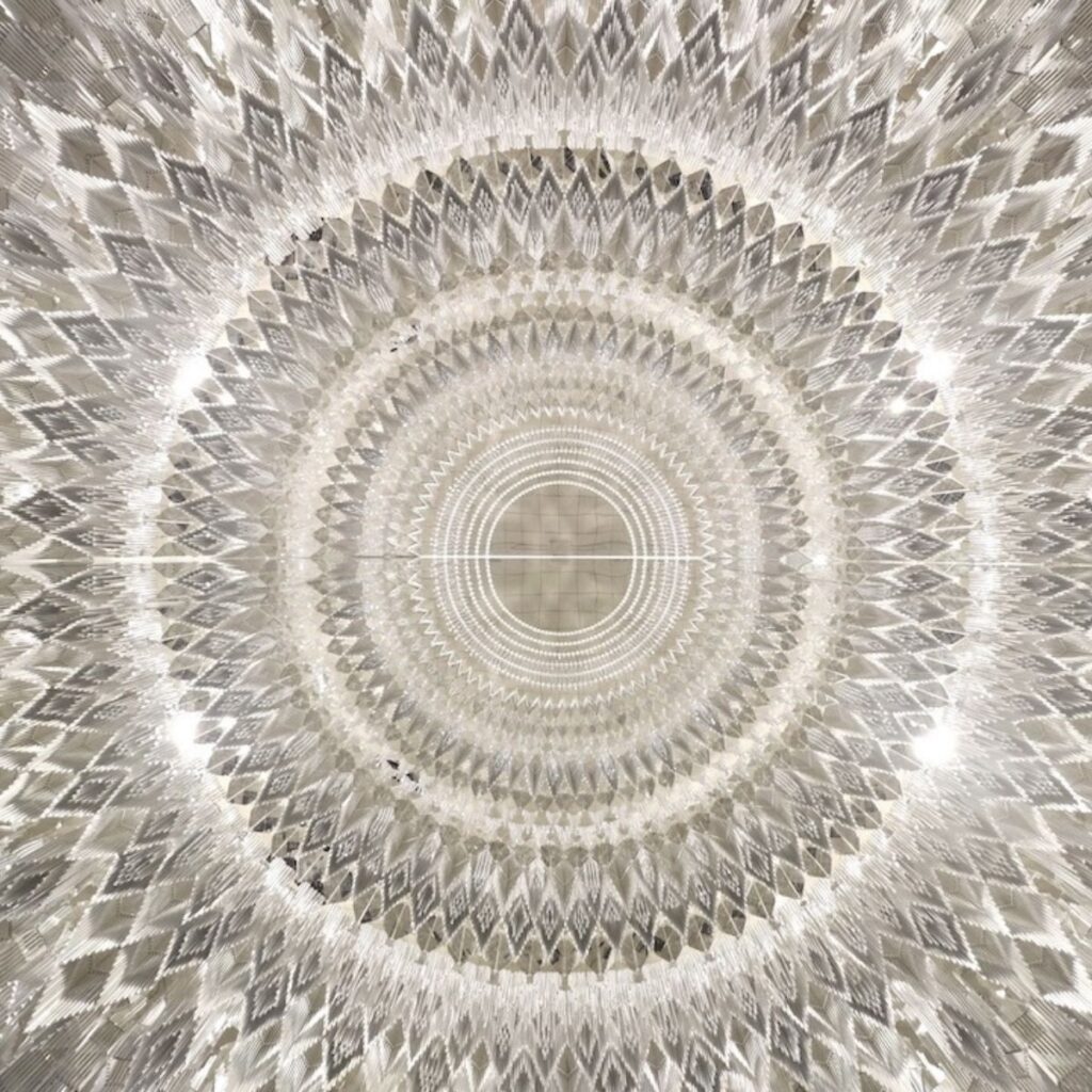 Looking up at a circular, geometric ceiling design with intricate diamond patterns radiating outward, creating a symmetrical, kaleidoscopic effect in shades of white and silver.
