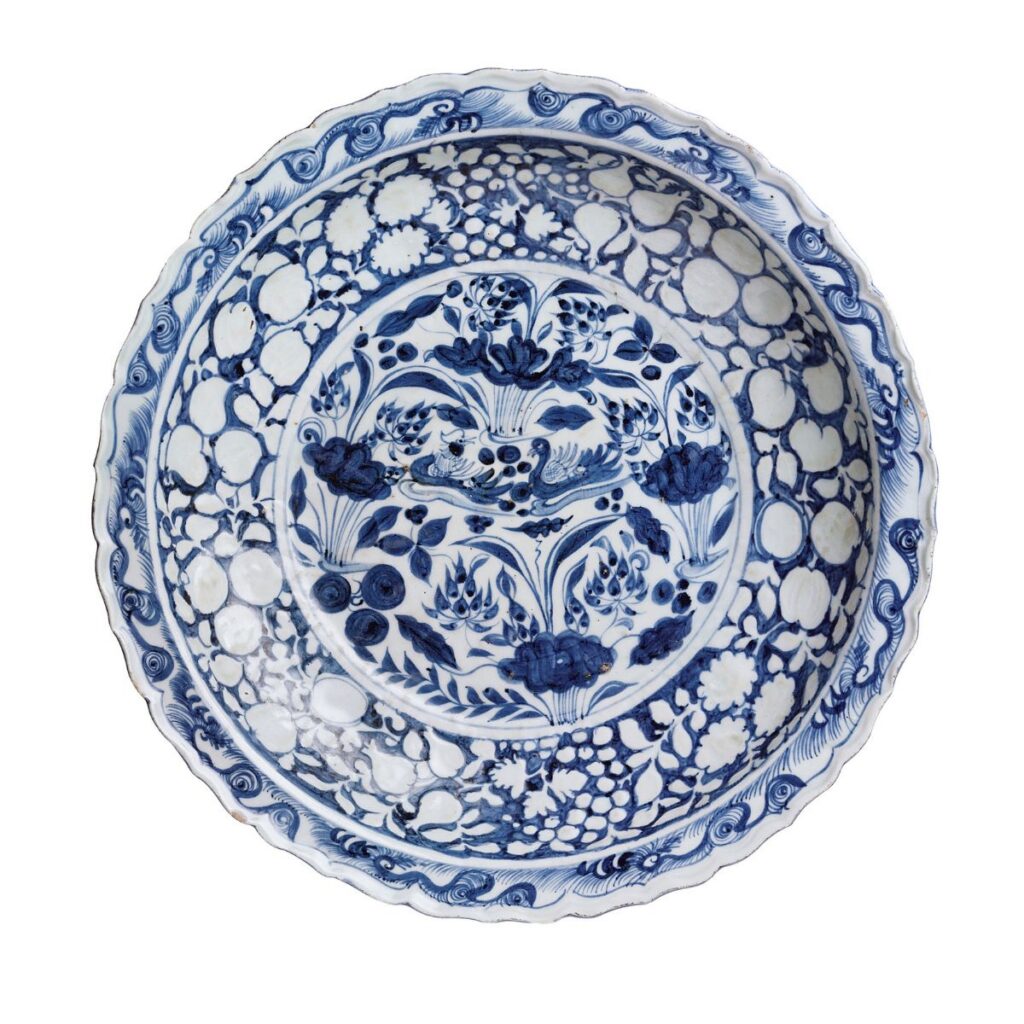 Round ceramic plate decorated with blue and white floral patterns reminiscent of boats floating afar, featuring central birds surrounded by a wide scalloped rim.