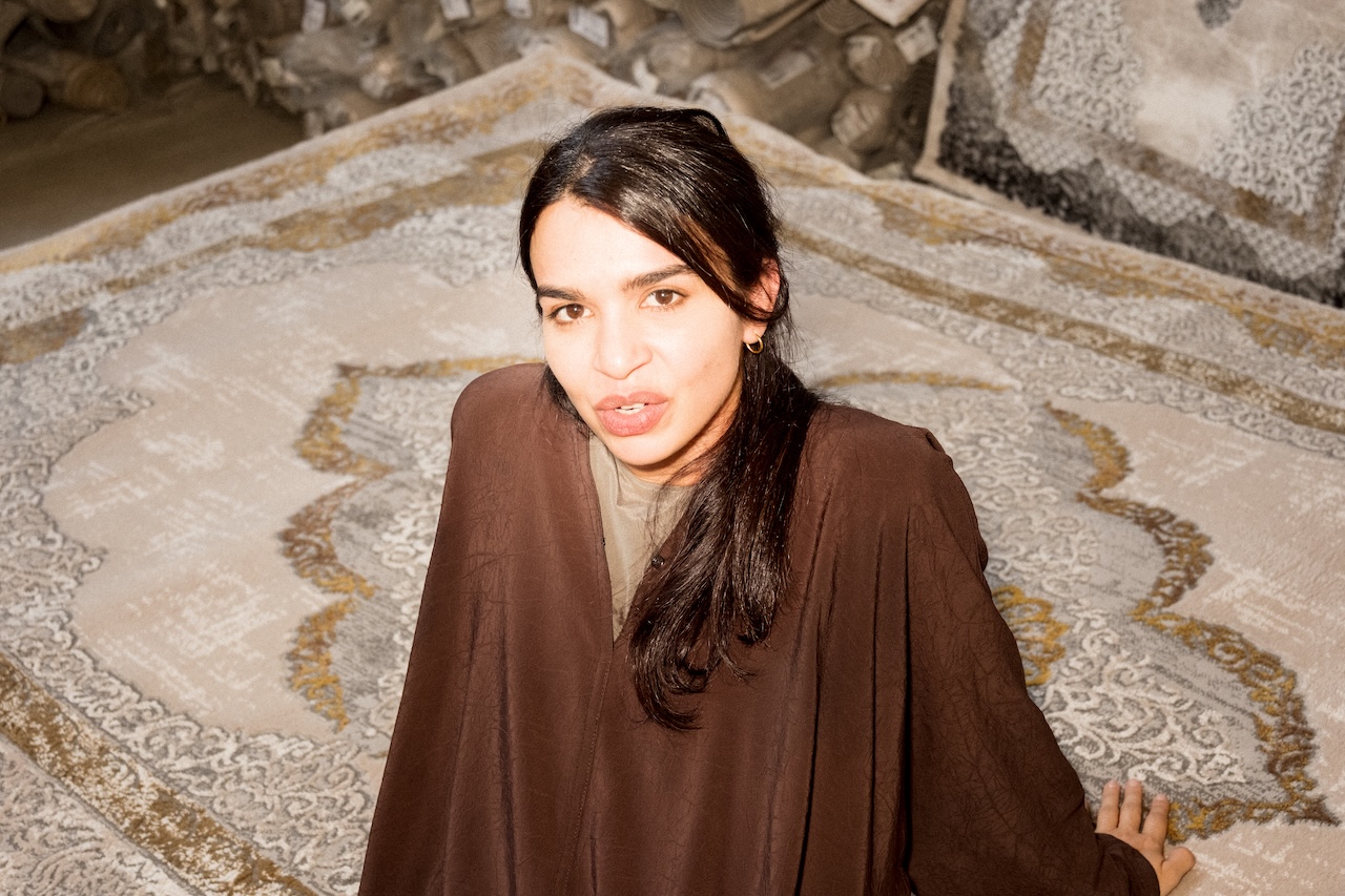 A person with long dark hair, wearing a brown outfit, sits on a patterned rug with rolled carpets in the background