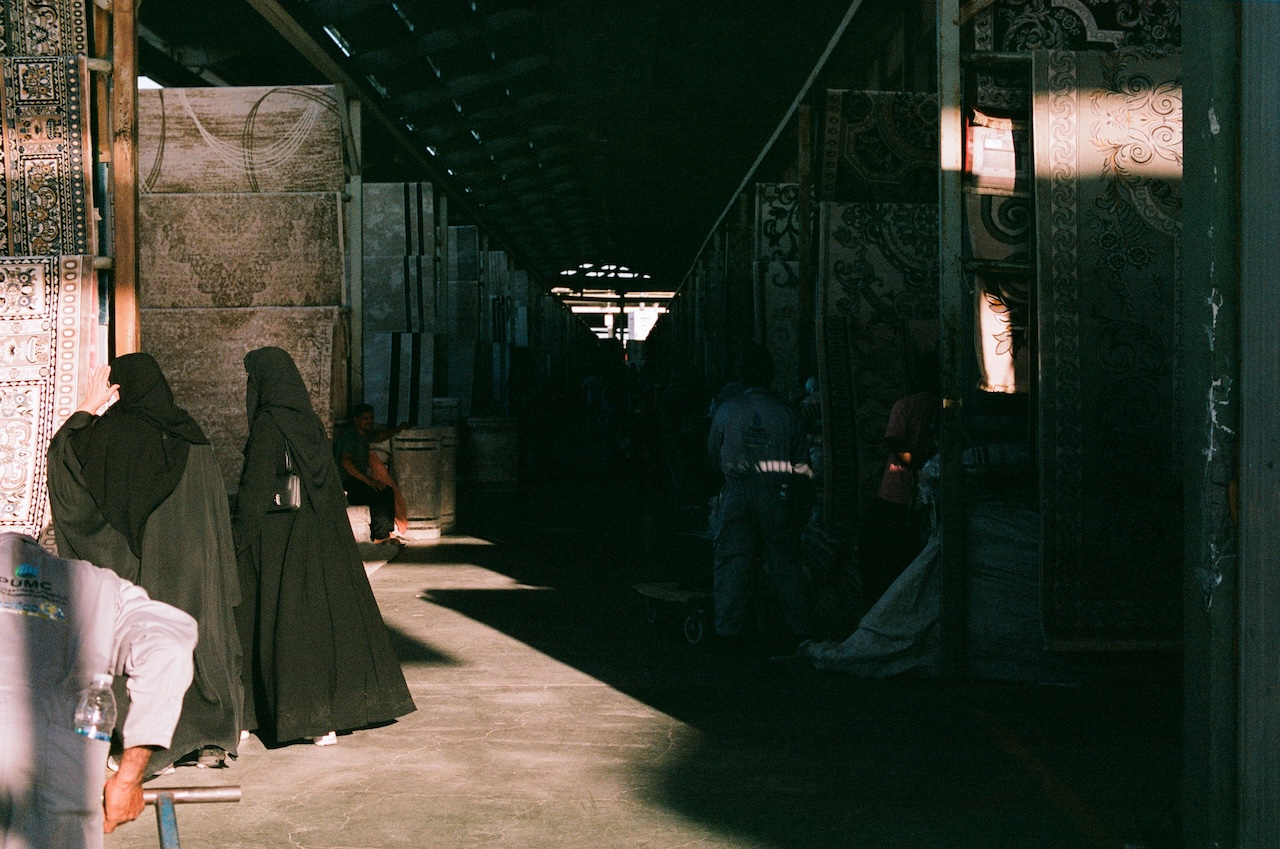 People walk through a dimly lit market corridor lined with large hanging carpets. Beams of sunlight highlight parts of the scene, casting dynamic shadows. Some people wear dark traditional clothing, adding to the atmospheric allure.