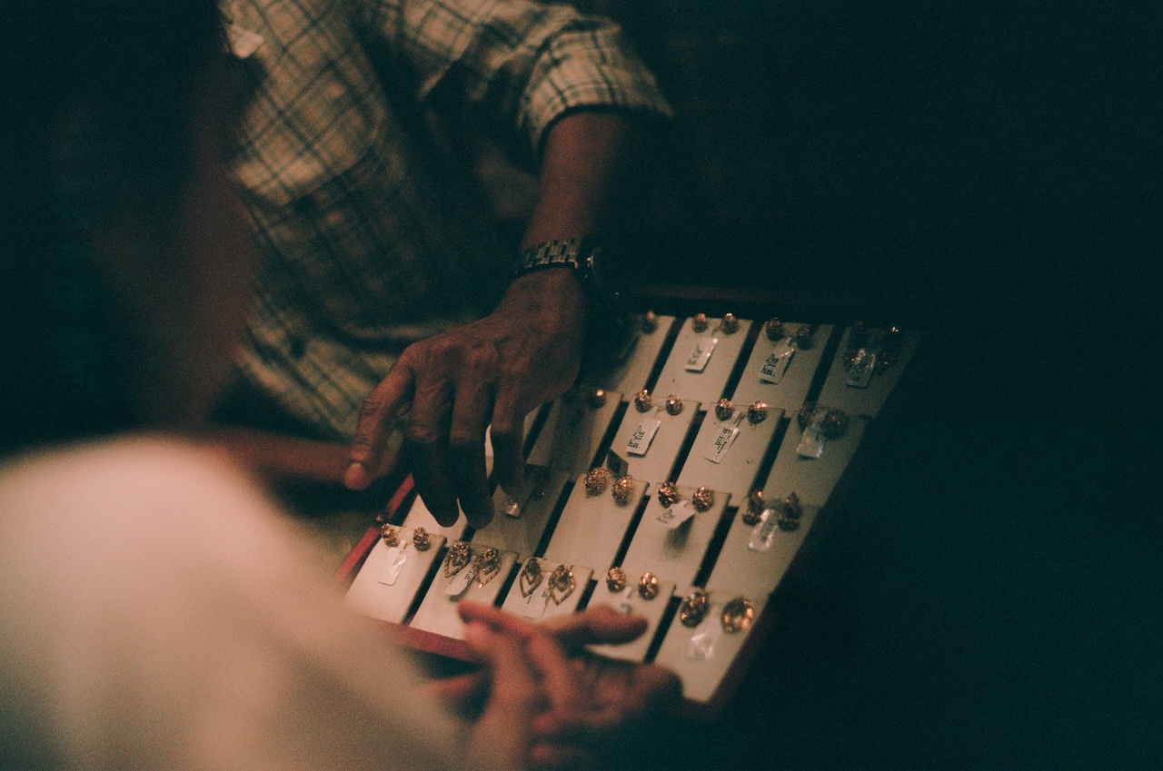 A person’s hand points at a display tray of various rings, possibly gold, while another person observes. The scene appears dimly lit, suggesting an intimate or indoor setting