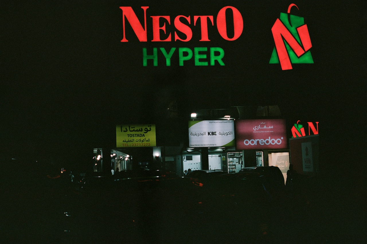 A night scene shows illuminated signs for Nesto Hyper in red and green above smaller storefront signs, including Ooredoo, KBE, and one in Arabic. Dark vehicles are parked in front, lending an urban atmosphere