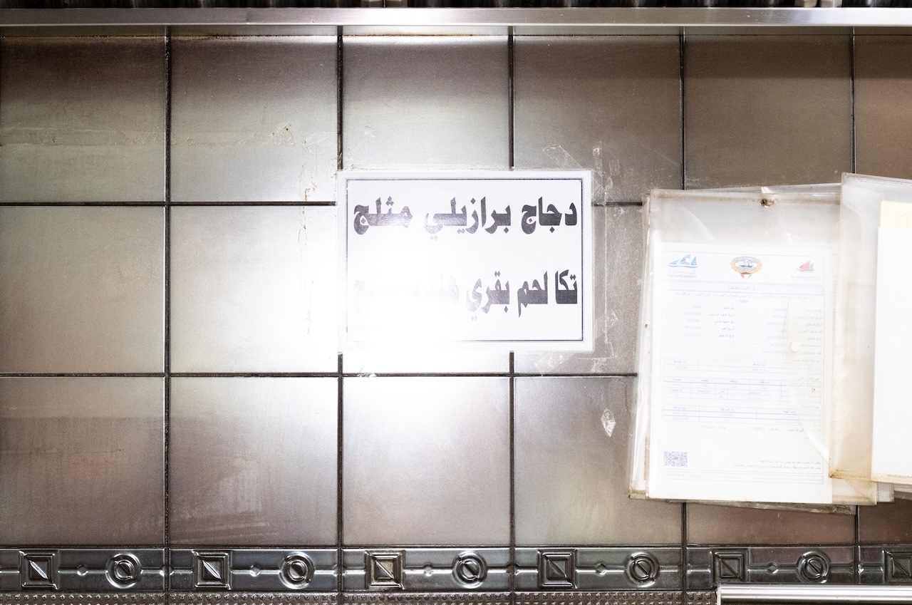 A tiled wall with a sign in Arabic above some papers. The sign is partially obscured by glare, making it hard to read clearly. The area appears clean and industrial, possibly part of a kitchen or food service area