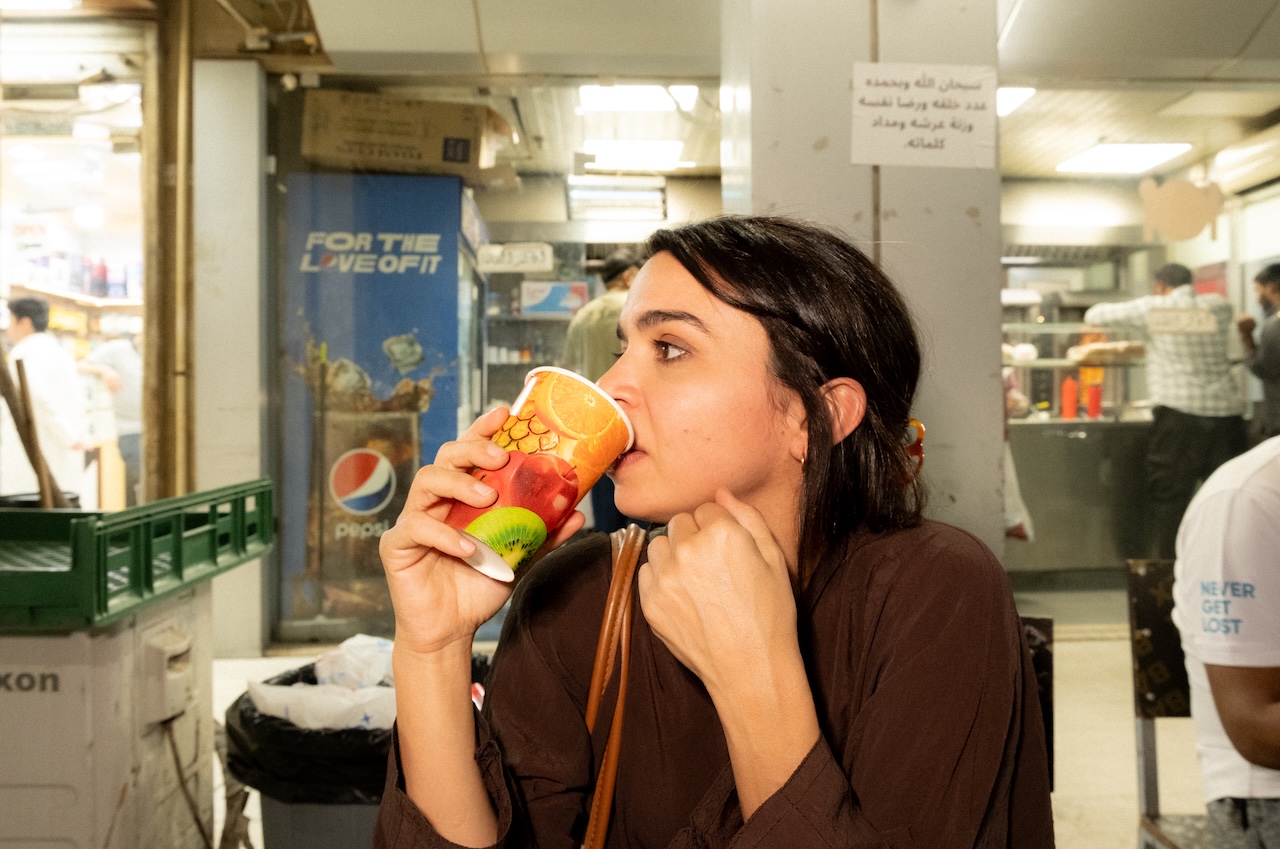A woman with dark hair sips a drink from a colorful cup with fruit images, sitting at an outdoor food stall at night. The background shows a Pepsi sign and people working in a shop