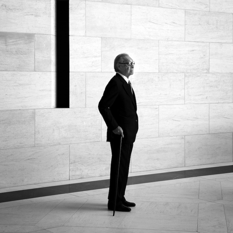 An elderly man in a suit stands indoors, holding a cane and looking to the left.