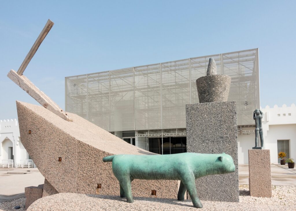 A bronze-colored sculpture of a cat by Rirkrit Tiravanija stands before a large, abstract stone structure and a modern building with scaffolding.