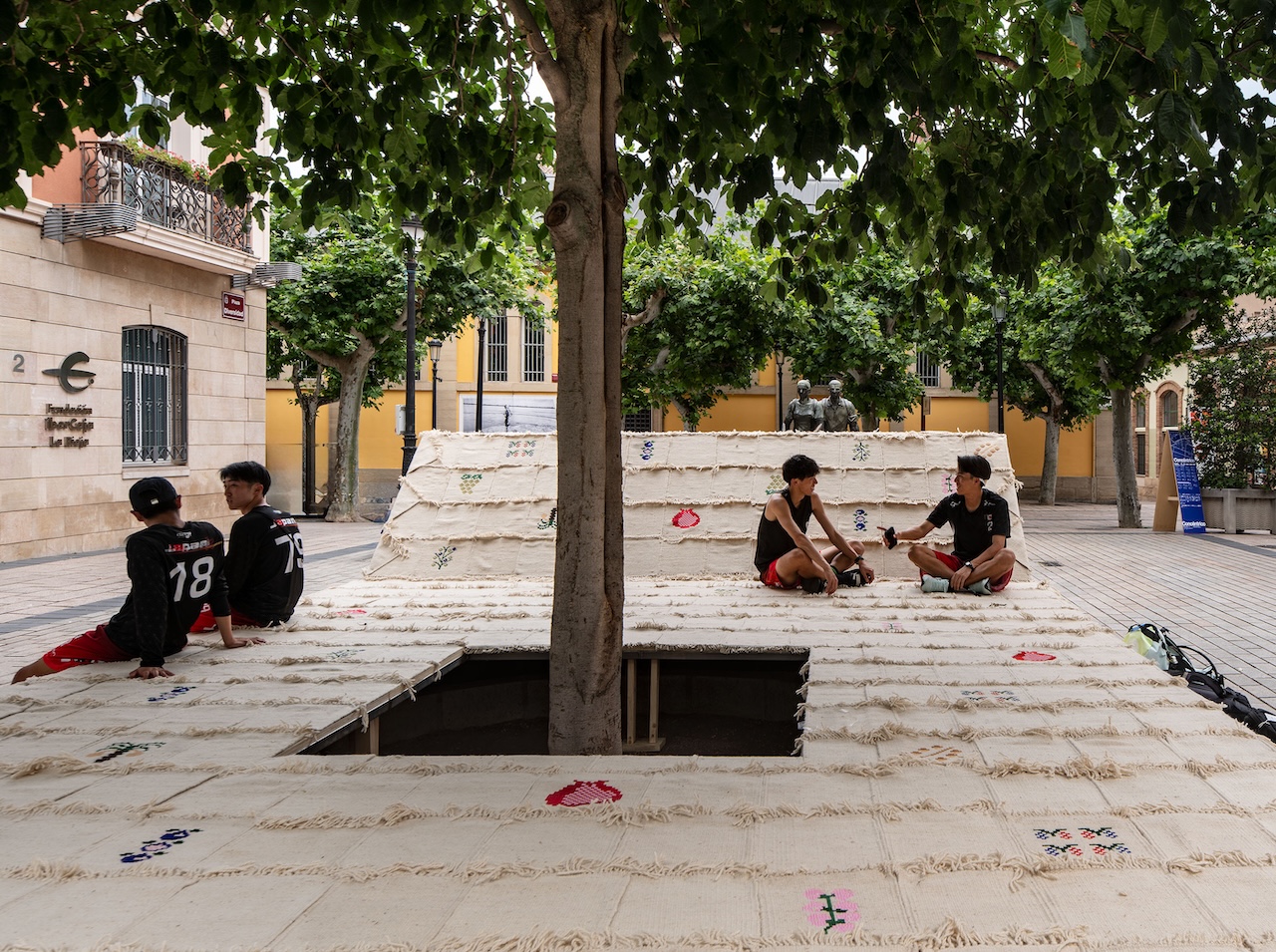 Four boys sit and talk on a large, elevated platform built around a tree in a city square. The platform is covered with textured white fabric and some colorful patterns, surrounded by buildings and more trees.