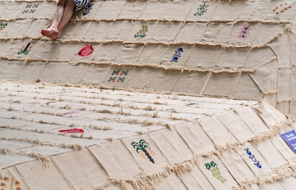 A childs bare feet rest on a large woven textile covered with small, colorful embroidered patterns and fringed edges. Only the legs and feet are visible.