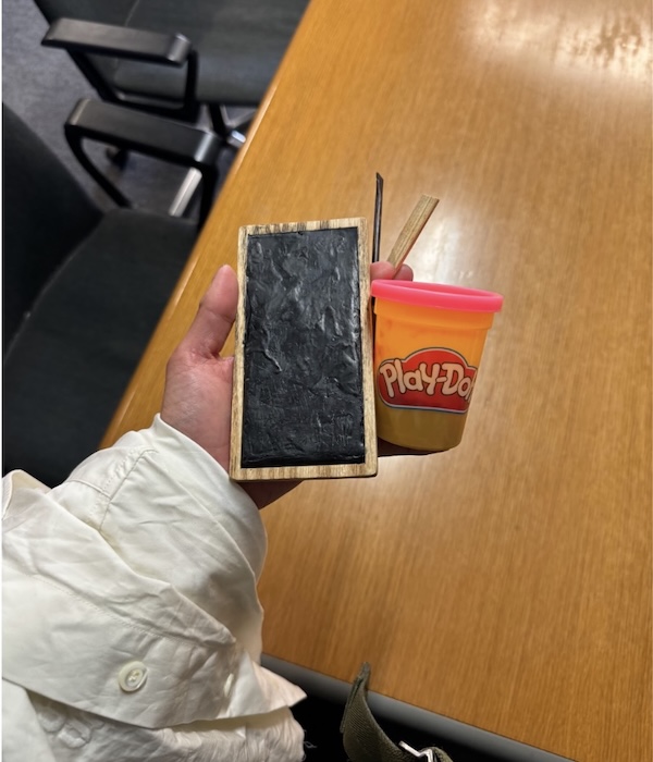 A person holds a small chalkboard, a stick of chalk, and a container of Play-Doh over a wooden table, with chairs visible in the background