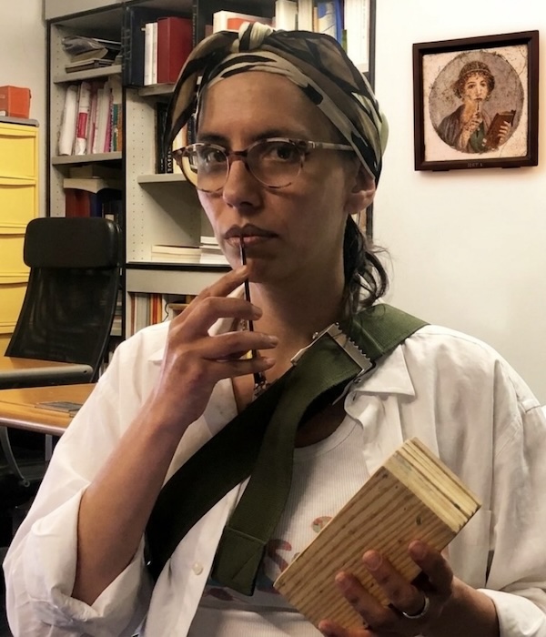 A woman wearing glasses and a headscarf holds a wooden object and pen, appearing thoughtful in an office with bookshelves and a framed artwork
