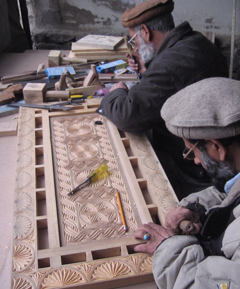 Luis Monreal and another man in flat caps carve an intricate geometric pattern into a large wooden panel.