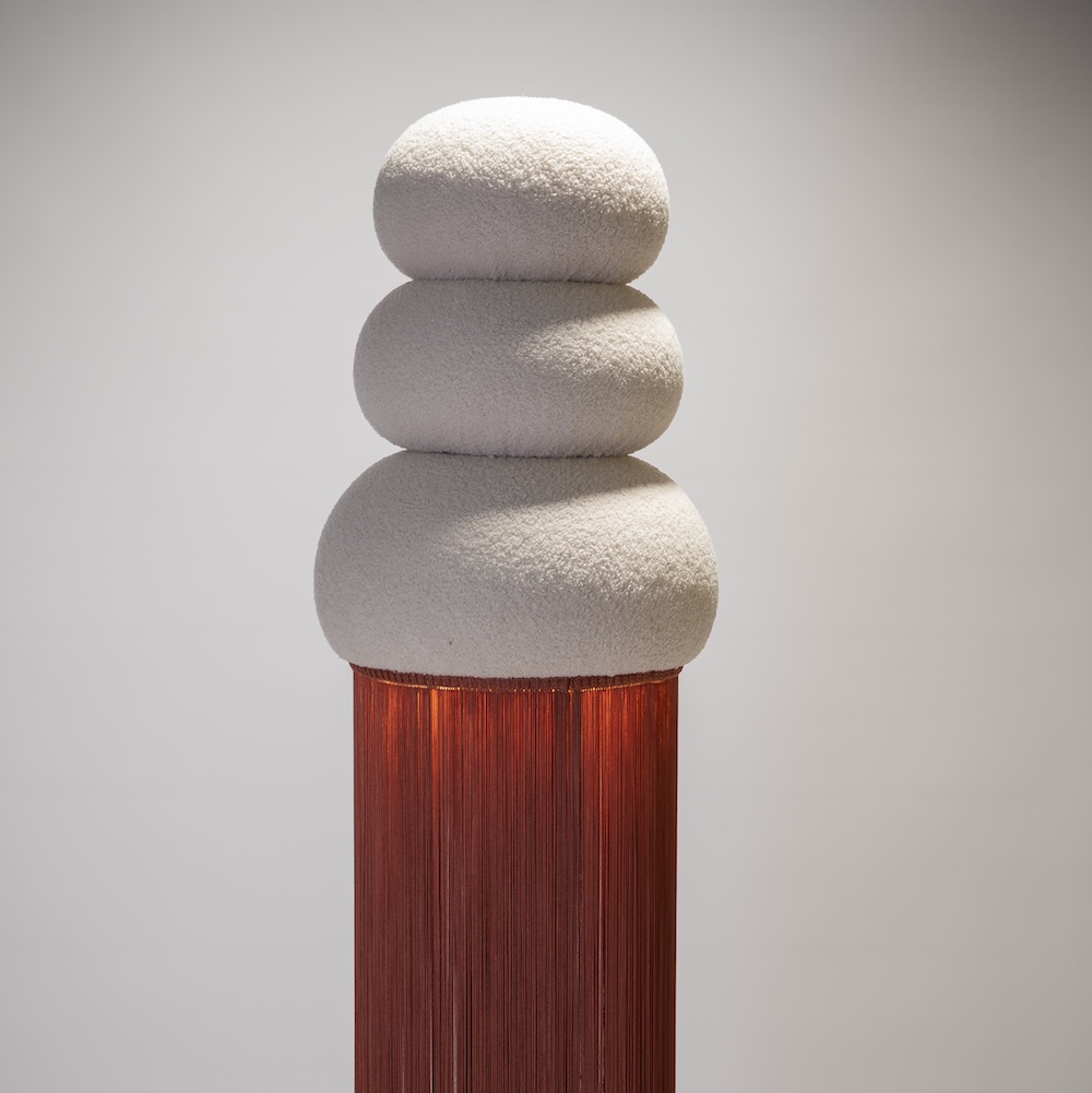 A sculpture features three smooth, white, stone-like shapes stacked on top, sitting above long, hanging red threads that cascade down. The background is a plain light gray.