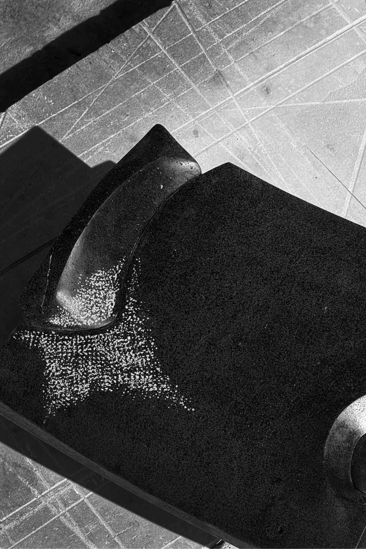 A close-up of a textured, curved black object with scattered white specks, casting shadows on a scratched, sunlit surface with intersecting lines