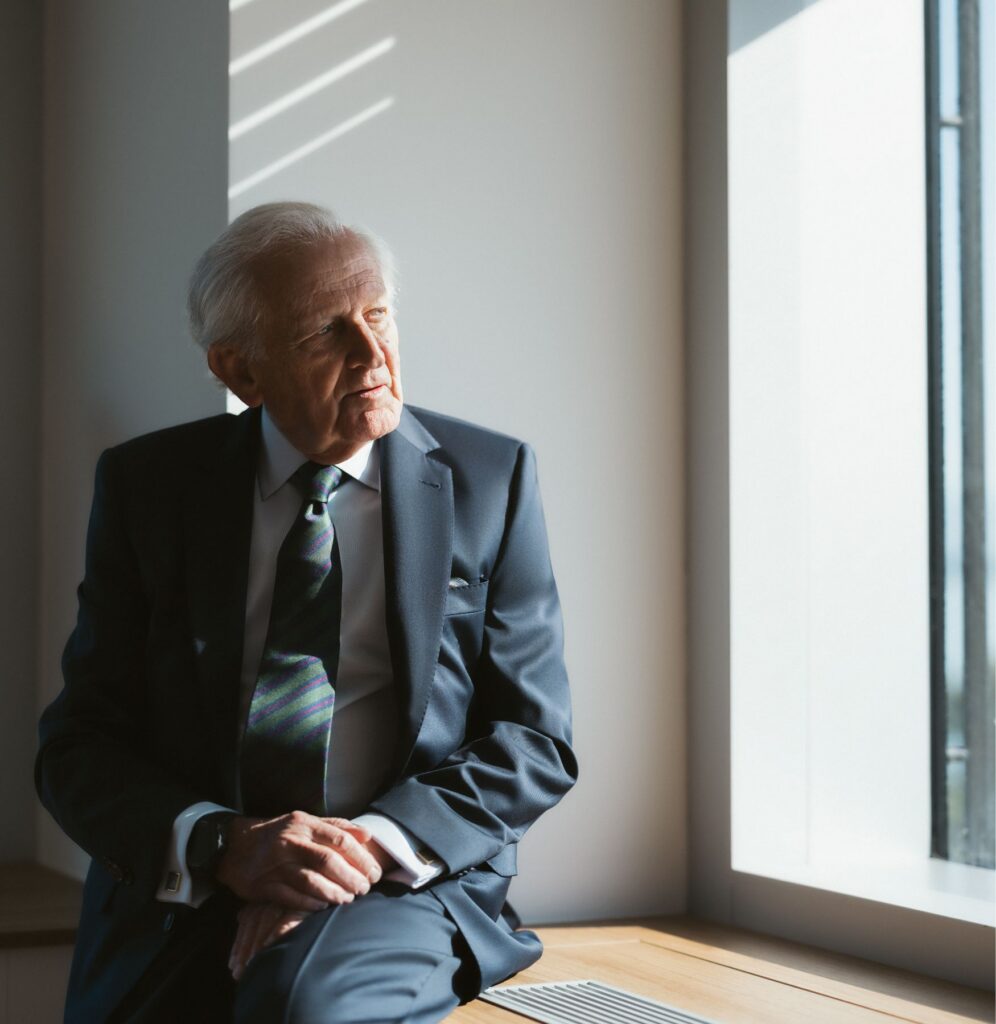An elderly man in a dark suit sits by a sunlit window, gazing thoughtfully outside with his hands resting in his lap, recalling memories of the Franca Fund Gala.