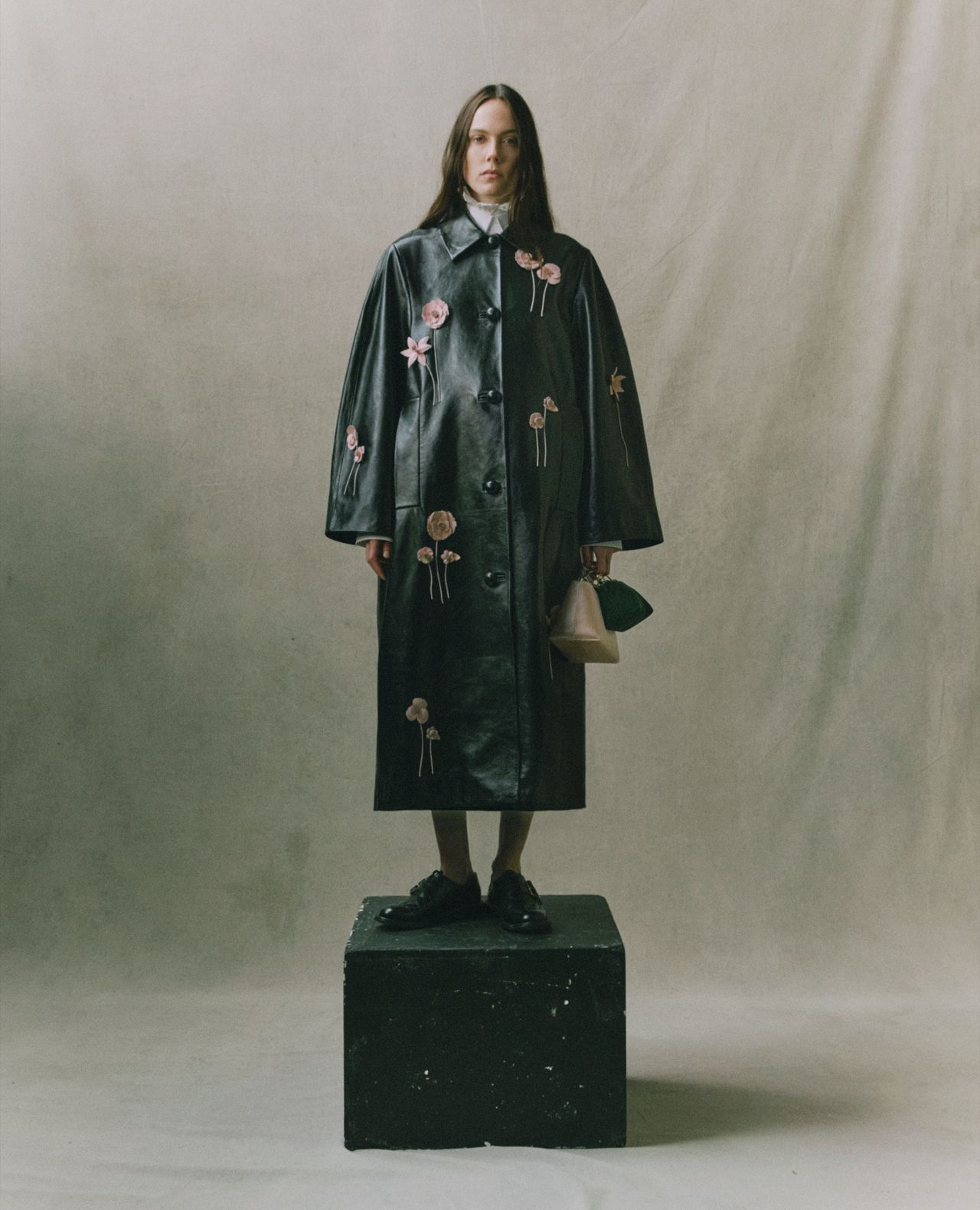 A person stands on a black box wearing a long, black leather coat with pink flower appliqués, black shoes, and holding a small beige bag