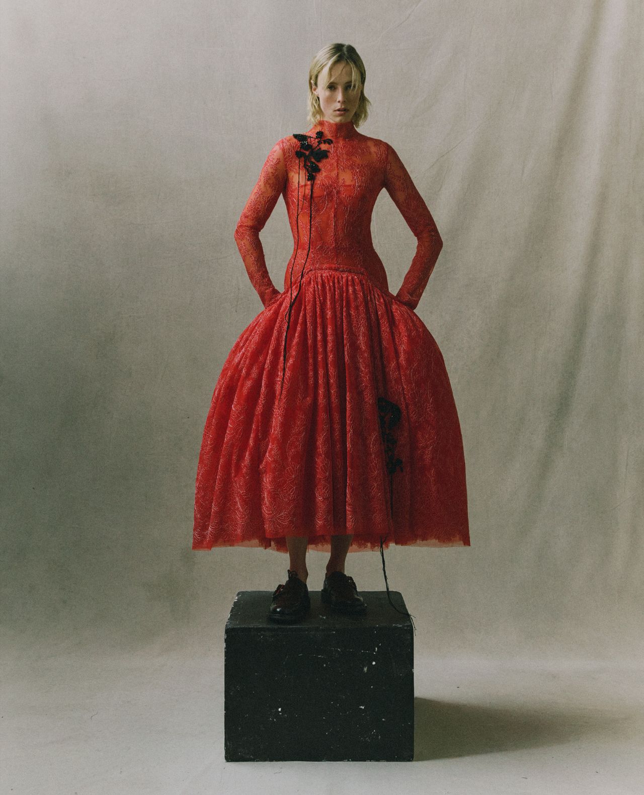 A woman stands confidently in a long, bright red lace dress atop a black box against a neutral backdrop. With hands on hips and black floral accessories draped across her dress and neck, she exudes both elegance and poise.