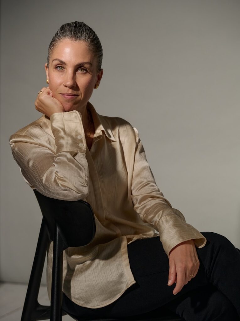A woman with slicked-back hair, wearing a shiny beige blouse and black pants, sits on a black chair resting her chin on her hand and smiling softly in a studio setting, reminiscent of an elegant portrait one might see at the Franca Fund Gala.