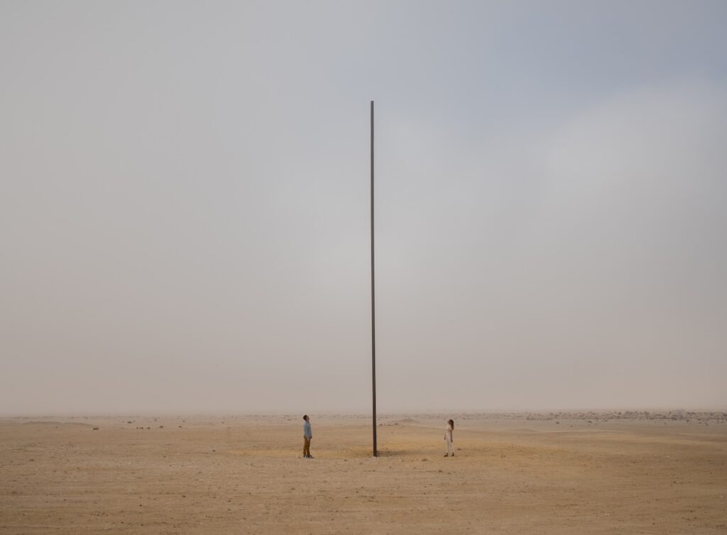 Two people stand on either side of a tall, thin vertical monolith by Rirkrit Tiravanija, set in the middle of a vast desert landscape.