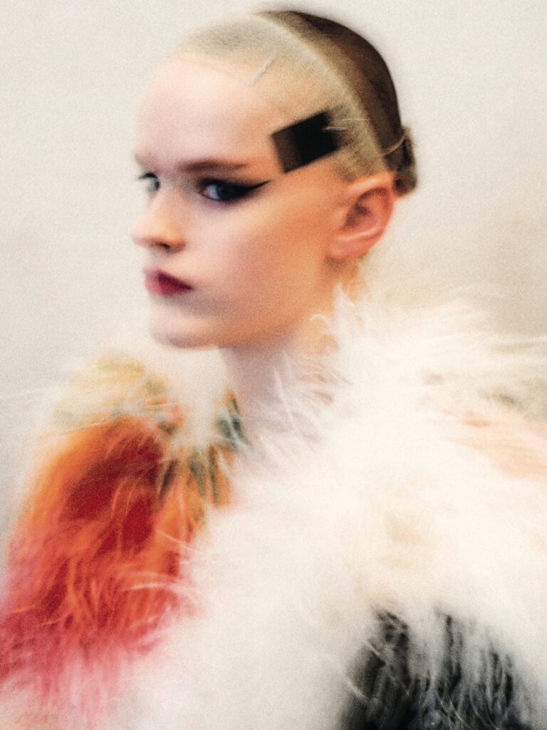 A person with slicked-back hair, bold black eyeliner, and a hair clip wears a fluffy, multicolored fur garment reminiscent of Chef Noof Al Mari’s avant-garde style. The image is softly blurred, creating an ethereal, dreamy effect.