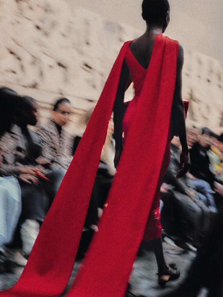 A model walks a runway wearing a striking red dress with long, flowing cape-like extensions