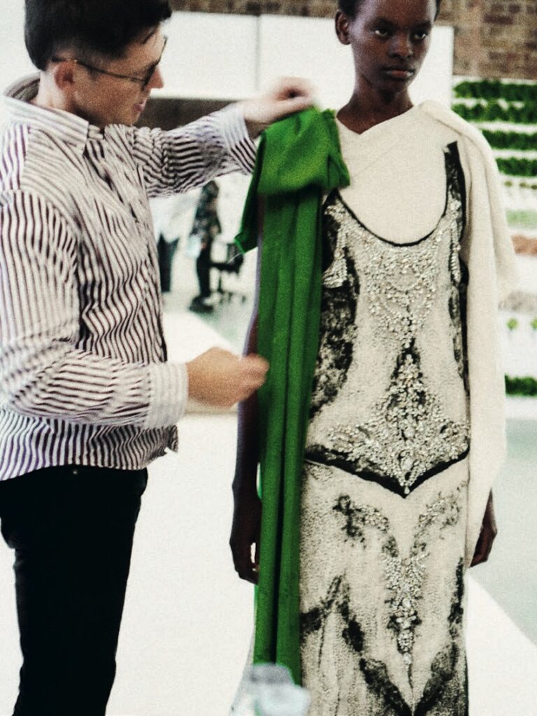 A person in a striped shirt adjusts a vibrant green fabric on a model wearing an ornate black and white dress