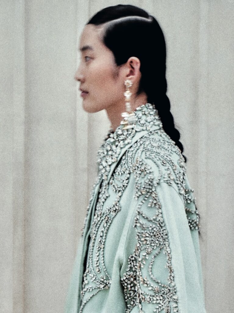 A person with a braided hairstyle wears a pale, embellished garment adorned with intricate beadwork and large dangling earrings, facing left against a soft, neutral background