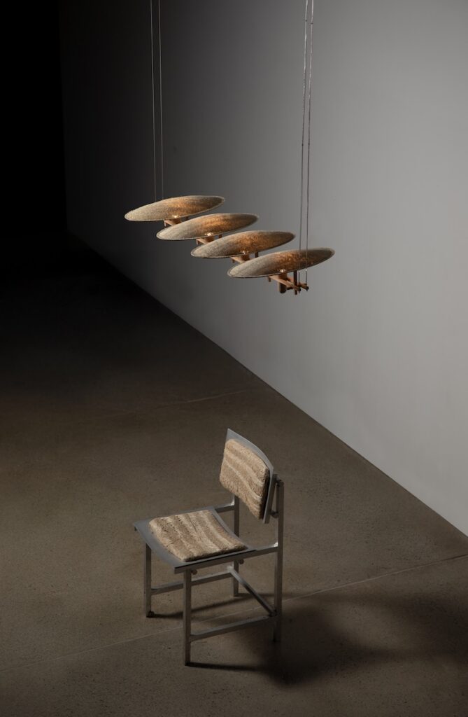 A metal chair with textured brown padding sits beneath four surfboard-shaped objects suspended from the ceiling in a dimly lit room