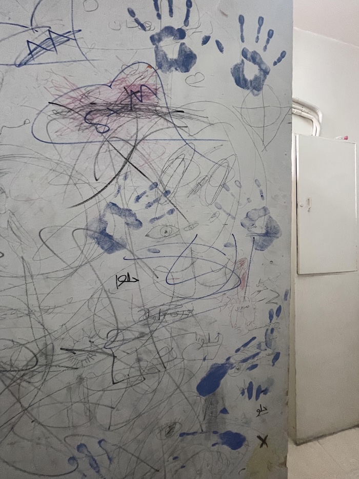 A wall covered in scribbles, drawings, and blue handprints, with a heart shape and the word SAM written inside it
