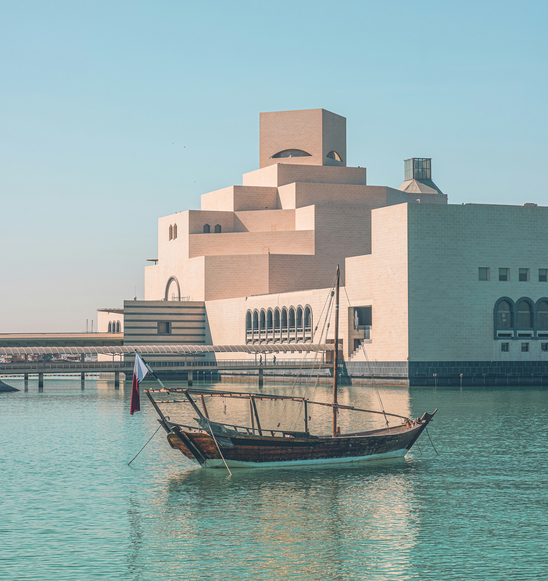 A traditional wooden boat floats on calm water near the modern geometric Museum of Islamic Art in Doha, Qatar, under a clear blue sky