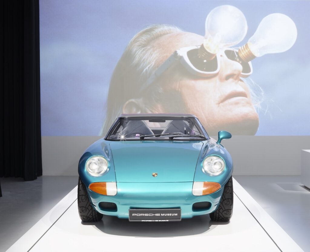 A turquoise Porsche sports car on a display platform in front of a large Rirkrit Tiravanija projection of a man wearing lightbulb glasses.