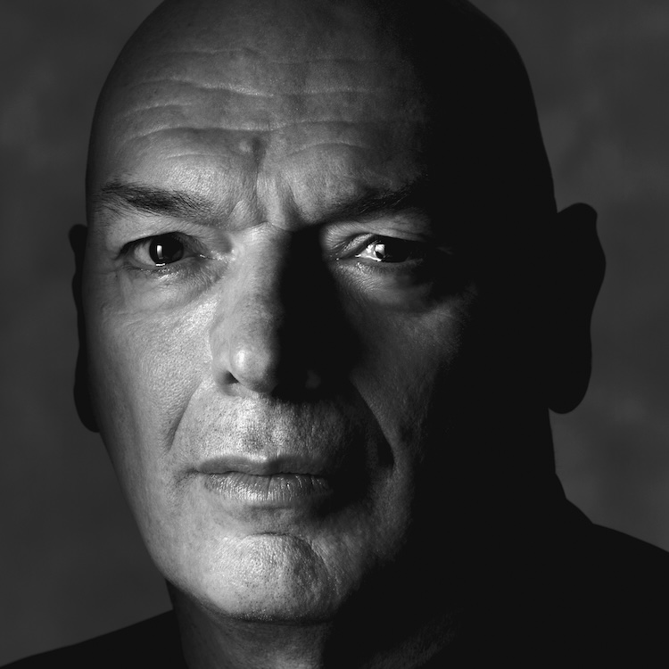 Black and white close-up portrait of the bald artist Rirkrit Tiravanija looking directly at the camera.