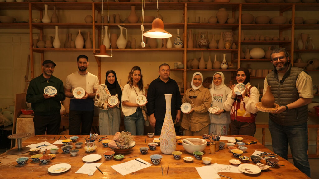 Nine people stand behind a long wooden table in a Qatar pottery studio, each holding a ceramic plate or bowl, with shelves filled with various ceramic vessels in the background.