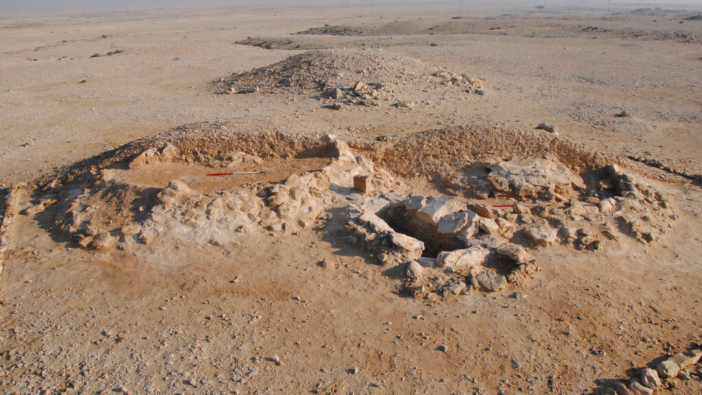 An archaeological site with stone ruins and a sunken stone chamber in the vast, arid desert landscape of Qatar.