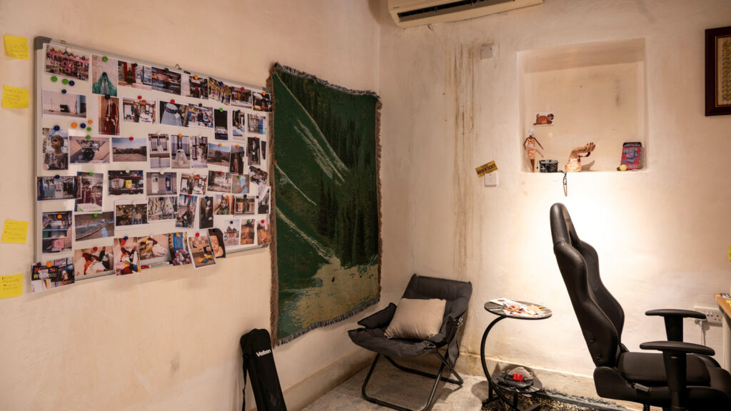 A room with a Qatar landscape wall hanging, a large photo collage pinned to the wall, a folding lounge chair with a pillow, and a black office chair near a small table.