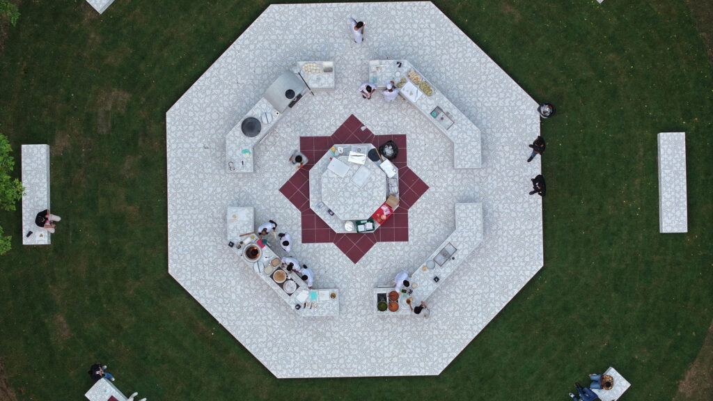 Aerial view of a hexagonal outdoor kitchen in Qatar with counters arranged around a central station, where people prepare and serve food on a patterned tile patio set in a grassy area.