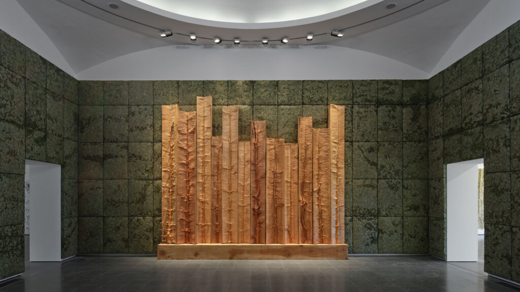 A large sculpture in Qatar made of vertical wooden planks with rough, uneven edges centered on a wall covered in dark, textured square panels.