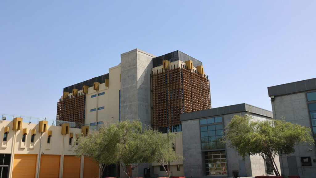 Modern multi-story building in Qatar featuring sections of concrete, yellow panels, and decorative latticework, with trees in the foreground.