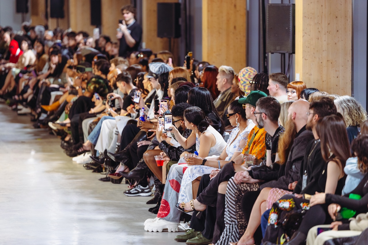 A row of people seated at a fashion show runway, many holding up smartphones to take photos.